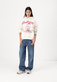 Cream hoodie with graphic print featuring a sun and mountain scene, paired with blue baggy jeans and light-colored sneakers.