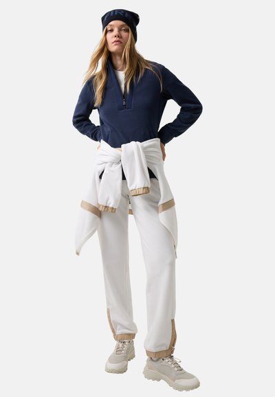 Marineblå fleece-pullover med lynlås, hvide joggers med brune accenter, indpakket hvid hættetrøje og beige sneakers med teksturerede såler.
