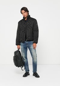 Sort quiltet pufferjakke med høj krave, parret med lyseblå slidte jeans. Modellen holder en sort rygsæk og har sorte sko på.