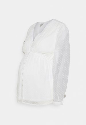 Blusa de maternidade branca com bolinhas texturizadas, fechamento por botão, decote em V e cintura franzida. Apresenta mangas longas e transparentes e um tecido suave.