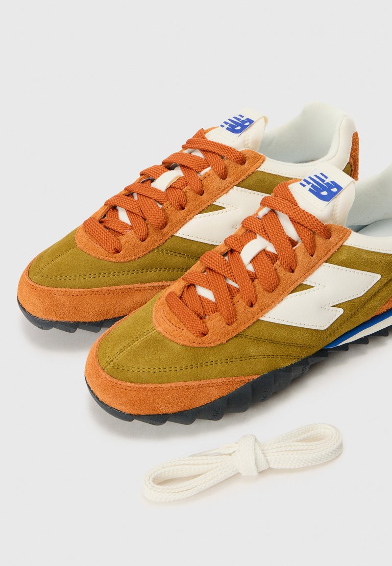 Un par de zapatillas de ante en verde oliva y naranja quemado con logo blanco, cordones naranjas y cordones blancos extra sobre un fondo blanco.