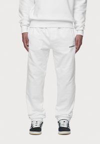 Bílé tepláky s elastickým pasem a manžetami, vybavené bočními kapsami a malým černým logem na levém stehně. Odolná tkanina z bavlněné směsi.