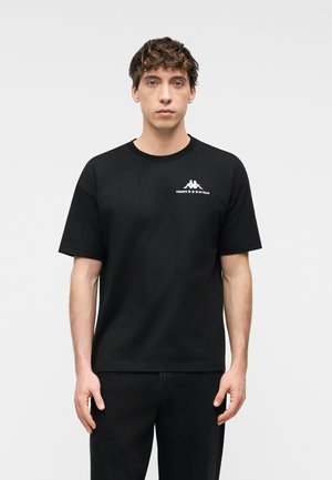 Νεαρός με σγουρά μαλλιά που φοράει μαύρο T-shirt με μικρό λευκό λογότυπο και κείμενο στο πάνω αριστερό μέρος του στήθους.