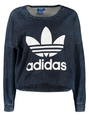 Persoon die een marineblauwe Adidas-trui, witte broek, zwarte Adidas sneakers draagt en een kleine zwarte schoudertas bij zich heeft.