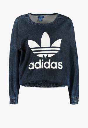 Personne portant un sweat-shirt Adidas bleu marine, un pantalon blanc, des baskets Adidas noires et un petit sac à bandoulière noir.