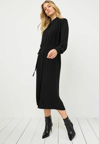 Czarna sukienka z długim rękawem, uszyta z miękkiego materiału, z kołnierzykiem na guziki i paskiem w talii. Czarne botki za kostkę z wąskimi czubkami i zamkami błyskawicznymi.