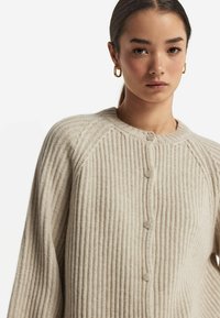 Cardigan beige à côtes avec un col rond fermé, doté de cinq boutons sur le devant et de longues manches amples en matériau doux et texturé.