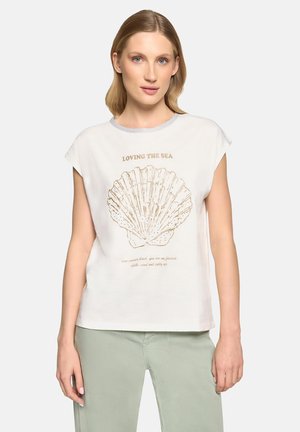 Frau trägt weißen ärmellosen Top mit grauem Saum, der eine große goldene Muschel und den Schriftzug „Loving the Sea“ zeigt, kombiniert mit hellgrünen Hosen.