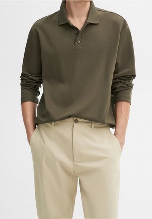 Mann trägt ein olivgrünes langärmeliges Poloshirt, in beige Hose gesteckt, mit den Händen in den Taschen vor einfarbigem Hintergrund.