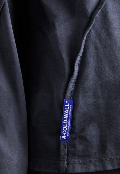 Svart stoffskjorte med synlig søm og en blå etikett med "A-COLD-WALL*" og teksten "BRITISH INDUSTRIAL SPORTSWEAR". Glatt tekstur.