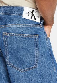 Denim bikses vidējā zilā krāsā ar kontrastējošām šuvēm un baltu Calvin Klein etiķeti uz jostas. Iezīmēta aizmugurējā kabata ar redzamām šuvēm.
