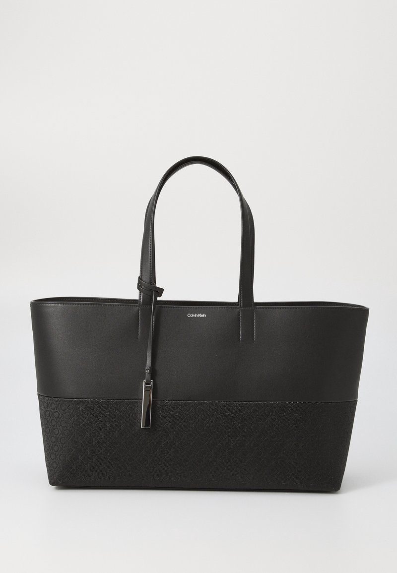 Černá taška přes rameno s hladkou horní částí a texturovaným dnem, které má jemný monogramový vzor. Úzké uši a dekorativní táhlo zipu.