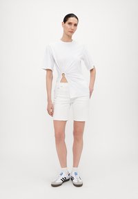 Hvit t-skjorte med korte ermer og vridd frontdesign som har en sirkulær åpning, kombinert med hvite shorts og svart-hvite sneakers.