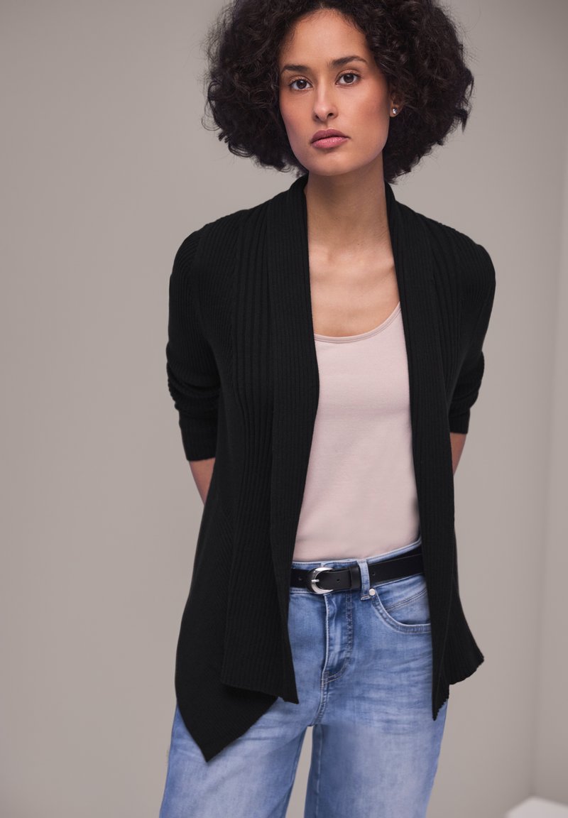 Frau mit lockigem Haar, die eine schwarze offene Strickjacke, ein blassrosa Oberteil und blaue Jeans mit einem schwarzen Gürtel trägt, vor einem schlichten Hintergrund steht.