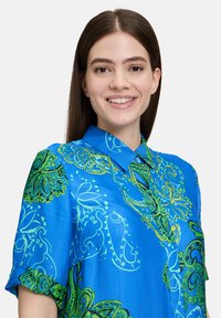 Camicia blu a maniche corte con dettagli intricati in paisley verde, tessuto liscio e design del colletto classico.