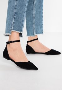 Chaussures plates en daim noir avec bout pointu, dotées d'une fine bride à la cheville et d'un petit talon, associées à un jean bleu clair à ourlet effiloché.