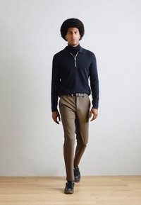 Mørkeblå langermet polo-skjorte, beige bukser og svarte joggesko. Skjorten har glidelås krage. Tregulv og nøytral vegg som bakgrunn.