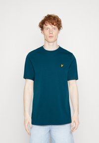 Camiseta de algodón de manga corta en color verde azulado, con escote redondo y un pequeño logo amarillo en el lado izquierdo del pecho. Textura suave.