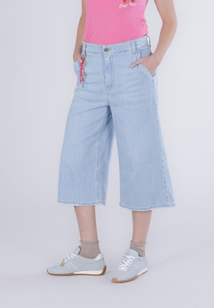 Osoba nosí světle modré široké džíny zkrácené na kotnících, růžový top, béžové ponožky a světle modré tenisky se šňůrkami a přívěskem.