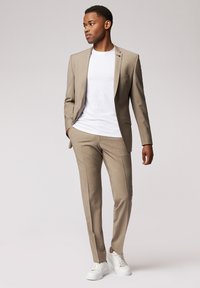 Beige Anzug mit einem einreihigen Sakko und schmal geschnittenen Hosen, kombiniert mit einem weißen T-Shirt und weißen Sneakers. Glatte Textur.