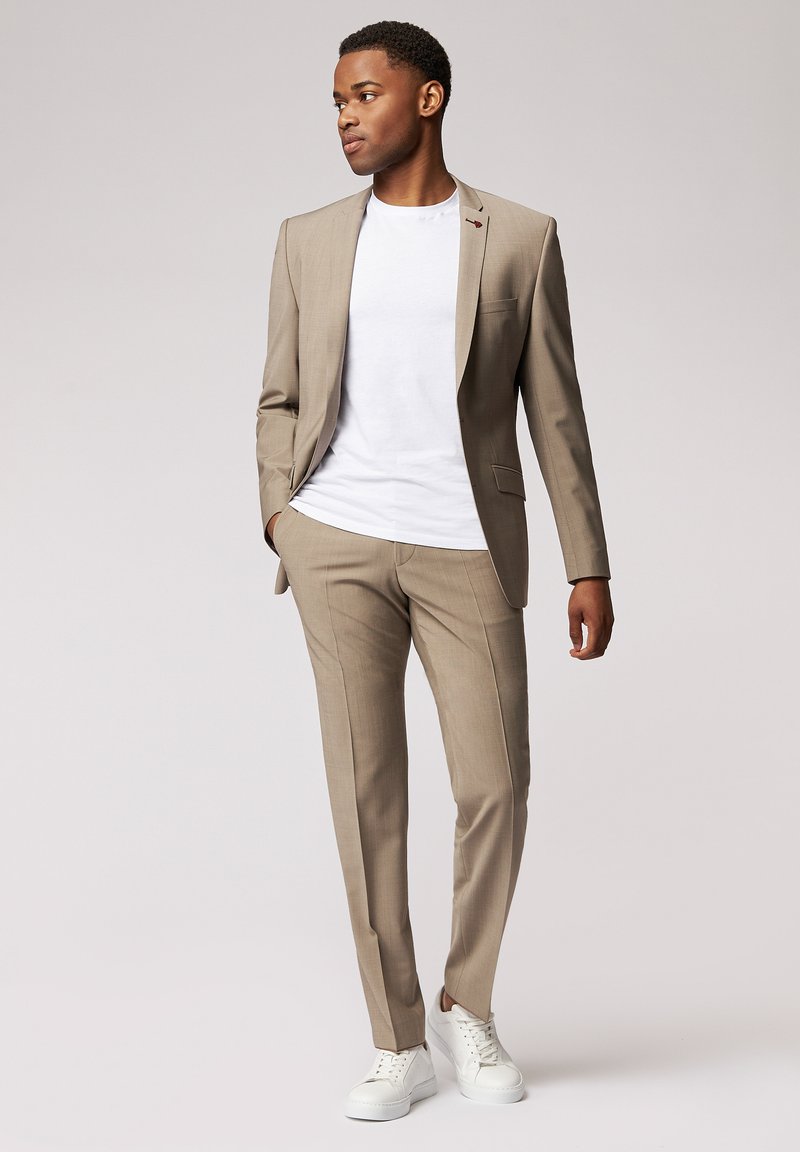 Beige Anzug mit einem einreihigen Sakko und schmal geschnittenen Hosen, kombiniert mit einem weißen T-Shirt und weißen Sneakers. Glatte Textur.