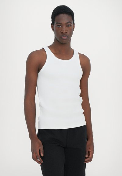 Débardeur blanc à côtes avec larges bretelles, coupe près du corps et texture lisse, porté avec un pantalon sombre. Style simple et polyvalent.