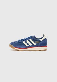 Blue suede sneaker with three white stripes, red accent on the sole, and a textured fabric near the ankle. Gold logo detail on side.