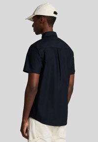 Camicia nera a maniche corte con piega sul retro, colletto strutturato e vestibilità rilassata. Indossata con un cappellino color crema. Design semplice e minimalista.