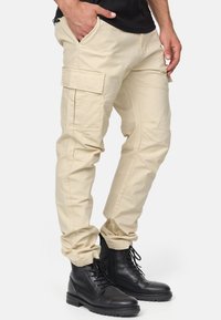 Pantaloni cargo beige con più tasche, vestibilità affusolata e tessuto leggero, abbinati a stivali neri stringati. Design semplice e funzionale.
