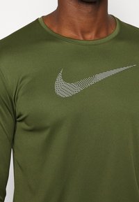 Mann trägt ein olivgrünes Langarmhemd mit einem weißen, stilisierten Nike-Swoosh-Logo auf der Brust.