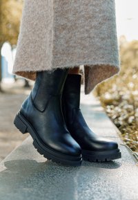 Botas de tobillo de cuero negro con un acabado suave, suela de goma gruesa y un diseño sencillo, complementadas con una falda beige de textura suave.
