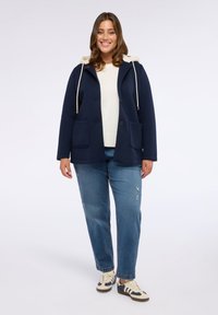 Marineblazer mit Kapuze, ausgestattet mit zwei Fronttaschen, Knopfverschluss und einem hellen Pullover darunter, kombiniert mit derben Jeans und Sneakers.
