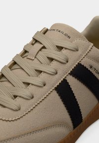 Baskets en suède beige avec des bandes en cuir noir, surface texturée, lacets plats et semelle en caoutchouc marron. Logo de la marque sur le côté.