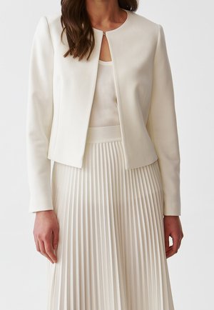 Kvinde iført en hvid jakke uden krave over en ærmeløs top kombineret med et hvidt, højtaljet, plisseret skørt på en lys baggrund.