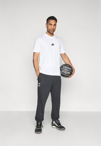 Camiseta blanca de manga corta de Adidas con logo negro, pantalones deportivos grises con logo lateral y zapatillas negras con detalles verdes, sosteniendo un balón de baloncesto.