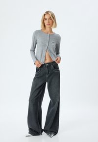 Grauer Langarm-Cardigan mit Knopfverschluss, kombiniert mit weit geschnittenen schwarzen Denim-Jeans. Verfügt über eine offene Front und eine glatte Stoffstruktur.