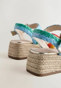 Sandały na platformie z plecionymi niebiesko-zielonymi paskami i masywnymi koturnami o fakturze sznura na jasnym tle.