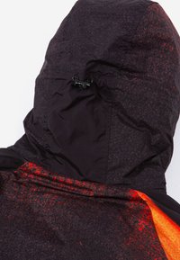 Mētelis ar kapuci, kuram ir melna pamatne un spilgti oranžs-sarkans akcenti. Tam ir teksturēts audums un regulējama aukla kapuces daļā.