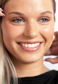 Gros plan sur une personne souriante avec des cheveux blonds clairs et des yeux bleus. La peau semble lisse et illuminée, avec un piercing au nez et un maquillage léger.