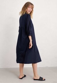 Tamsiai mėlynas, iki kelių ilgio suknelė su pūstomis rankovėmis ir laisvu siluetu. Audinys turi lygų paviršių. Dėvima su juodais austo stiliaus šlepetėmis.