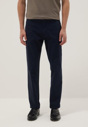 Man draagt nauwsluitende marineblauwe broek, zwarte loafers en een taupekleurig overhemd ingelegd, staat voor een effen lichte achtergrond.