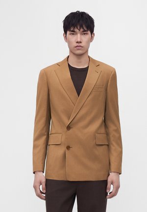 Ung mand med kort sort hår iført en camel-farvet dobbeltradet blazer over en mørk skjorte og mørke bukser mod en ensfarvet lys baggrund.