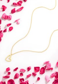 Goldene Kettenhalskette mit einem herzförmigen Anhänger, platziert auf verstreuten rosa Rosenblättern vor einem weißen Hintergrund.
