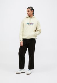 Beige hoodie met een grafische print, een voorzak en koordjes, gecombineerd met donkere corduroy broeken en witte sneakers.