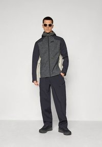 Grå och svart zip-up jacka med huva, med kontrasterande sidopaneler, kombinerad med mörka cargobyxor och stövlar. Solglasögon fulländar looken.