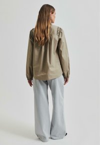 Kaki spalvos ilgos rankovės marškiniai su pūstomis pečių dalimis ir raukiniais, derinami su šviesiai mėlynomis plačiomis kelnėmis, matomi iš nugaros.