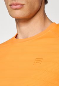 Suure plaan lähedalt kellegi seljas, kes kannab oranži tekstuuriga spordipluusi, mille rinnal on väike, reljeefne "F" logo.