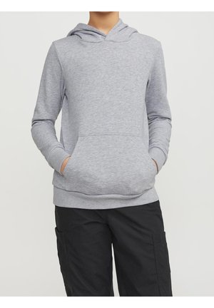 Person iført en lysegrå hættetrøje med frontlommer og sorte cargobukser, stående mod en ensfarvet lys baggrund.