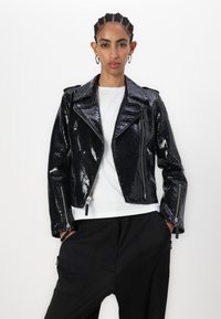 Veste de biker en cuir noir, brillant, au motif serpent, avec un col cranté, des zips argentés et des poches latérales, associée à un t-shirt blanc.
