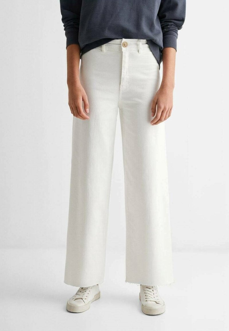 Jeans en denim blanc taille haute et coupe évasée avec un ourlet effiloché, doté d'une fermeture à un bouton et de poches avant. Associés à un haut foncé et des baskets.